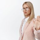 Serious young lady boss saying no, showing stop prohibition gesture, standing in suit and glasses, white background.